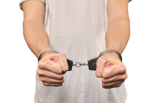 Hands of criminal in handcuffs, arrest of dangerous criminal isolated on white background, close up. Male fists locked in handcuffs, apprehension of offender in off-white T-shirt, violation of law
