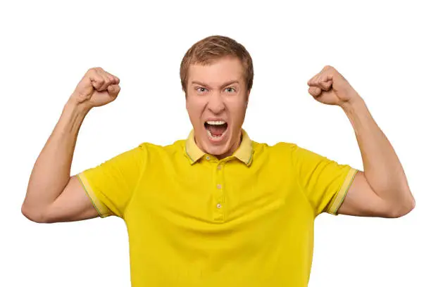 Funny young guy in casual yellow T-shirt raising hands and rejoicing of success isolated on white background. Excited crazy man showing muscles and shouting, joyful facial expression