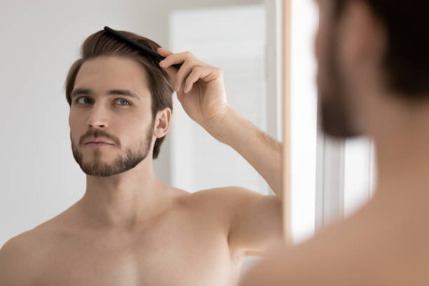 Handsome shirtless guy looks in mirror brushing his healthy hair Handsome shirtless bearded 30s guy standing in bathroom after shower looks in mirror holding wooden comb, brushing his healthy hair. Ad of male haircare cosmetics products, beauty treatment concept Styling Hair Gel stock pictures, royalty-free photos & images