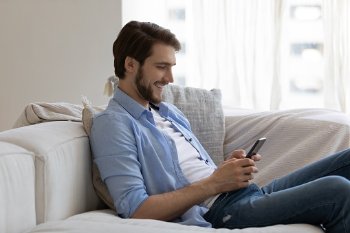 Smiling handsome millennial man sit on cozy sofa holding smartphone share messages through messenger. Handsome man buying on-line, enjoy easy web retail services. E-commerce and e-dating user concept