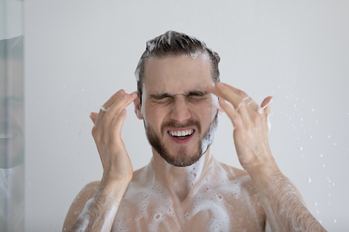 Head shot young lathered man takes a shower having eyes tearing up due to sulphate shampoo, haircare products, experiences soap allergy, frowns his face feeling discomfort and irritated, stinging eyes