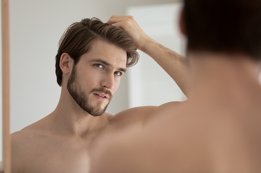 Young shirtless guy looks in mirror touch hair feels concerned due to receding, dry, dull hair. Hair loss common hair problem prevention, alopecia treatment ad, cosmetics products for male concept