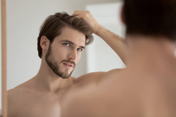 Man looks in mirror touch hair feels concerned due receding Young shirtless guy looks in mirror touch hair feels concerned due to receding, dry, dull hair. Hair loss common hair problem prevention, alopecia treatment ad, cosmetics products for male concept bald guys with beards stock pictures, royalty-free photos & images