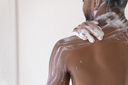 Cropped close up male body part, back and shoulder of African man take shower wash body use soap, hydrating gel, do morning self-hygiene routine. Skincare, bodycare product for men, healthcare concept