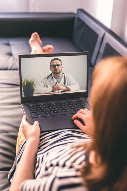 Cтоковое фото Женщина лежит на диване и разговаривает с врачом онлайн с помощью ноутбука. Концепция телемедицины.