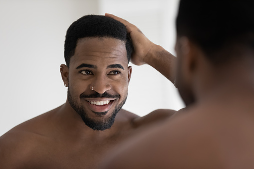 Handsome shirtless muscular build African man standing in bathroom looking in mirror touch his natural afro hair, concept of happy client of barber shop services, haircare treatment, cosmetics for men