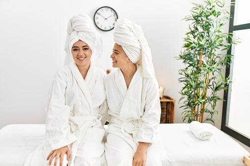 Woman couple wearing bathrobe hugging each other sitting on massage board at beauty center