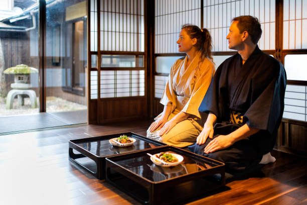 伝統的な日本の家庭の旅館の部屋に障子の引き戸のそばのテーブルプレートに座って着物焼座のカップル、石灯籠のある静かな庭を眺める - 日本食 ストックフォトと画像