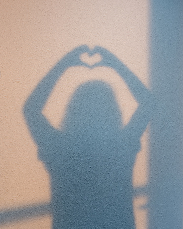 Young woman crating a heart-shaped shadow with her hands on a white textured wall