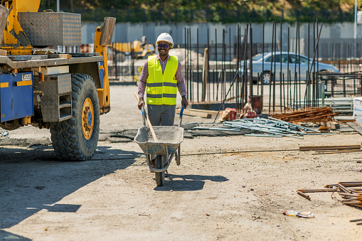 Black Man in overalls and a bright vest carries a wheelbarrow to pour building stone there for laying the road