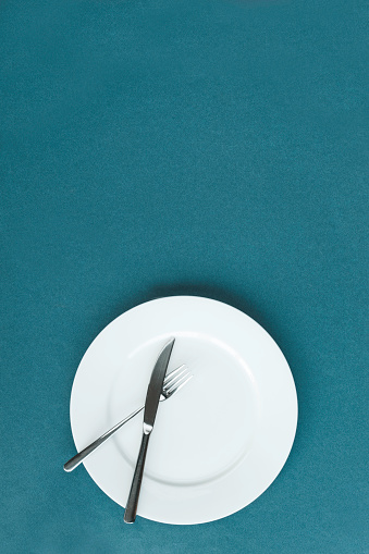 Directly above view of empty white plate, knife and fork on blue colored background. Representing celebration dinner.