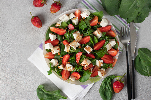 Healthy salad with strawberries, spinach, feta and pine nuts dressed with balsamic in plate on light gray background