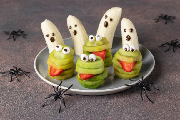 Spooky banana ghosts monsters and green kiwi monsters for Halloween party on brown background decorated with spiders, Halloween Fruit Serving Idea stock photo