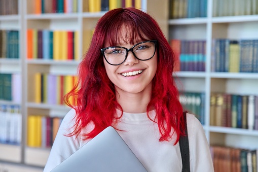 Portrait of female student with laptop backpack, looking at camera in library. Beautiful fashionable girl with dyed red hair wearing glasses, smiling with teeth. College, university, education, youth