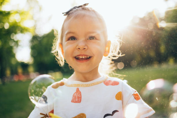 Portrait of a young excited girl making bubbles with a toy camera. She is outside in the park on a bright sunny day.
