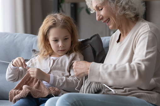 Smiling middle aged mature grandmother teaching adorable small child girl knitting stuff with needles and threads. Happy multigenerational female family involved in creative hobby activity at home.