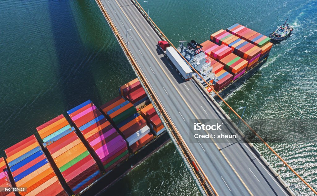 Aerial View of Container Ship Aerial drone view of a container ship navigating beneath a semi truck crossing a suspension bridge. Freight Transportation Stock Photo