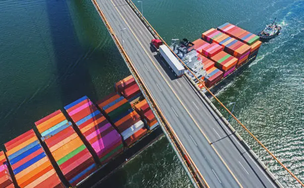 Aerial drone view of a container ship navigating beneath a semi truck crossing a suspension bridge.