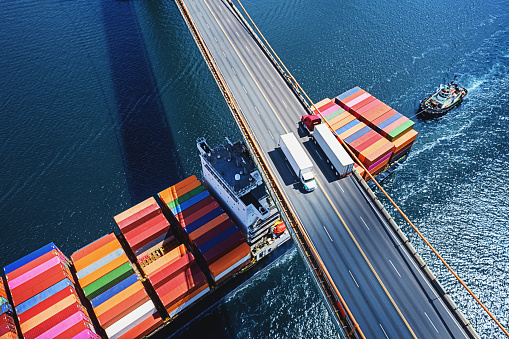 Aerial drone view of a container ship navigating beneath two trucks crossing a suspension bridge.