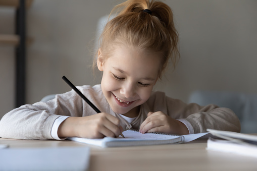 Head shot sincere happy small redhead child girl making notes in paper copybook, learning writing letters, preparing for examination or doing homework alone sitting at table, education concept.