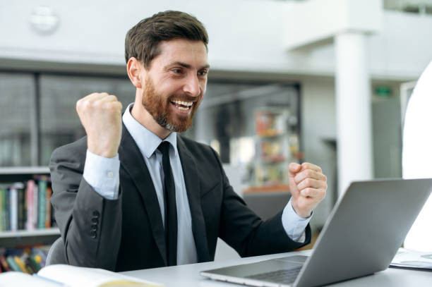 istock Positive joyful Caucasian businessman, web developer, IT specialist, programmer, sits and works at a laptop, in office, rejoices at the conclusion of a successful deal and the signing of a contract 1404543902
