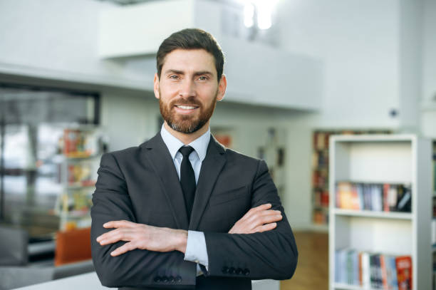 Confident attractive intelligent caucasian man, company director, office worker, top manager, wearing stylish business suit, standing in a modern office with arms crossed, looks at camera, smiling hombre caucásico inteligente y atractivo y confiado, director de la empresa, trabajador de oficina, gerente superior, vestido con un elegante traje de negocios, de pie en una oficina moderna con los brazos cruzados, mira a la cámara, sonríe