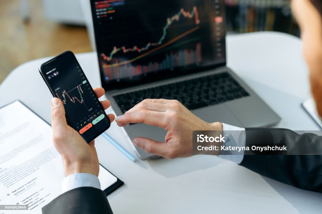 Successful male crypto trader, investor, using a laptop and smartphone, analyzes trading charts in the stock market of a digital cryptocurrency exchange, analyzes, buys and sells cryptocurrencies Stock Market and Exchange Stock Photo