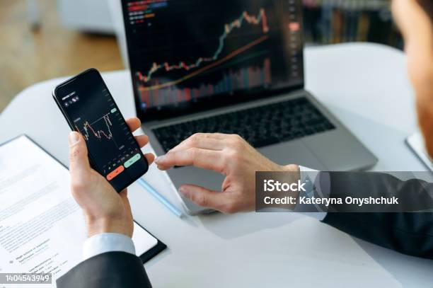 Successful Male Crypto Trader Investor Using A Laptop And Smartphone Analyzes Trading Charts In The Stock Market Of A Digital Cryptocurrency Exchange Analyzes Buys And Sells Cryptocurrencies Stock Photo - Download Image Now