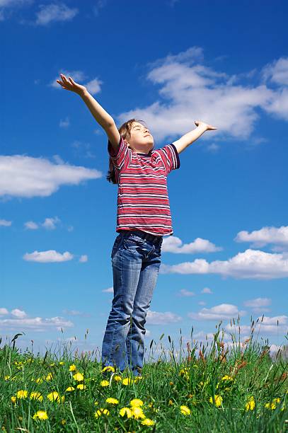 Young child standing in a field lifting arms toward the sky Young girl holding arms up against cloudy blue sky religion sunbeam one person children only stock pictures, royalty-free photos & images