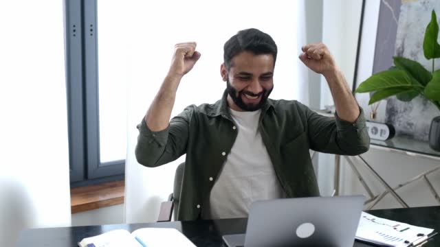 Happy excited lucky Indian or arabian man, business person, sits at a desk in modern office, got a good news, gesturing with his fists rejoicing in victory, success, good deal, big profit, smiling