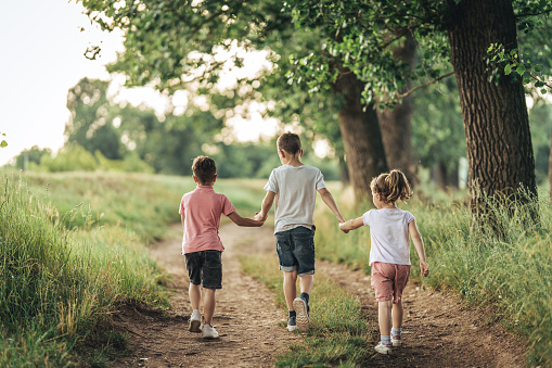 Two young boys and little girl holding hands all together while walking on dirt road in nature.