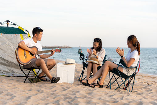 Happy friend have fun playing guitar and clap in camp they smiling together in holiday on sand beach near camping tent vacation time at sunset, Young Asian group woman and man in summer travel outdoor