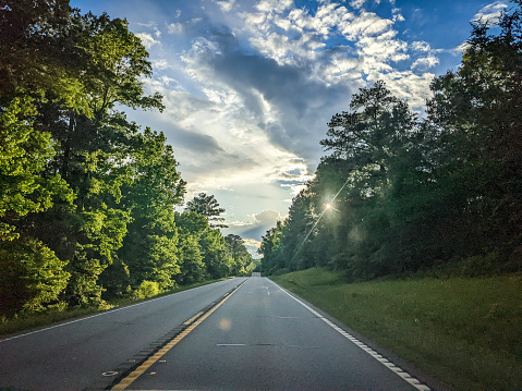 Horizontal photo from the perspective of driving down a tree-lined highway in Georgia. The sky is blue with white clouds and the sunlight is peeking through the trees.
