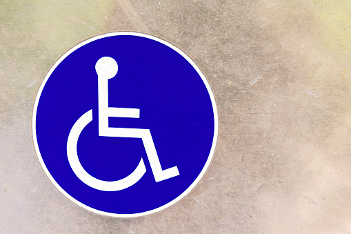 Close up of round blue sign with white symbol of person in wheelchair on textured background indicating person with disability