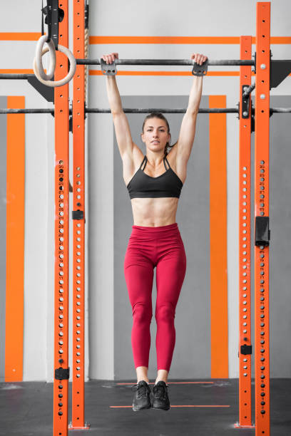Fit athletic woman doing a dead hang in a gym gym Fit athletic woman doing a dead hang from bars in a gym gym in a full length view for a healthy active lifestyle concept horizontal bar stock pictures, royalty-free photos & images