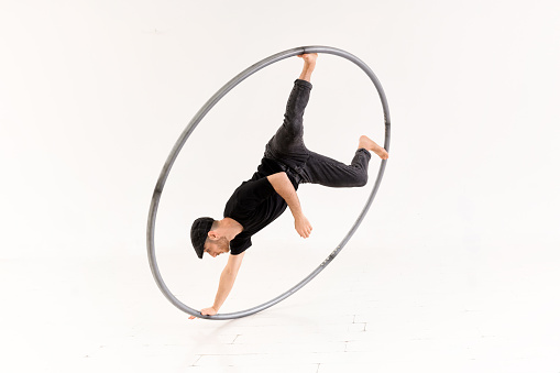 Full body of active barefoot acrobat performing one arm coin stunt with cyr wheel against white background in light studio