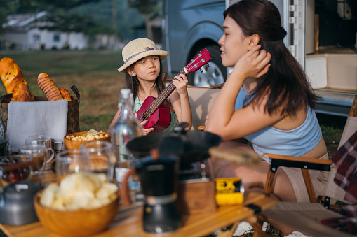 Happy Asian family having fun while little girl playing a guitar for her mother at picnic table in park.  Camper van travel series