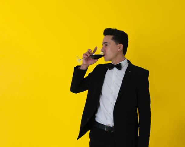 элегантный парень, одетый в галстук-бабочку, пьет чашку вина, стоящий впереди желтого цвета фона - bow looking at camera waist up indoors стоковые фото и изображения