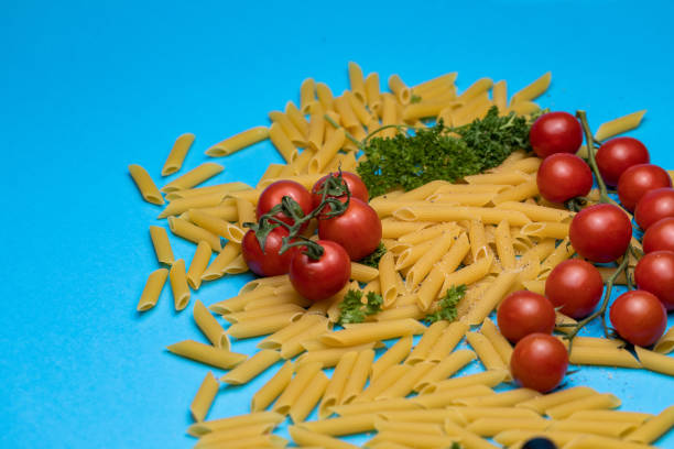 Close up shot of raw pasta tubes, cherry tomatoes and parsley on a bright blue background.