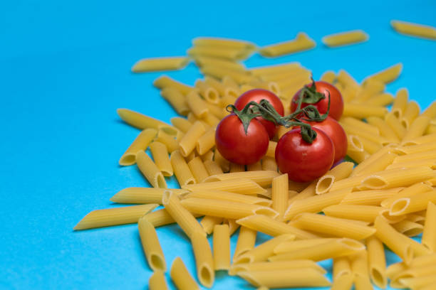 Close up of raw pasta tubes and cherry tomatoes on a bright blue background.