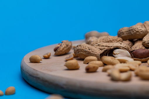 Close up shot of peanuts inside and outside of their shells on a cutting board on a bright blue background.