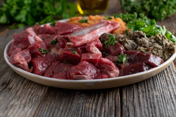 Plate with raw meat with beef muscle meat, rumen, offal, pureed vegetables, herbs and bones for healthy dog food. Served on a plate isolated on wooden table background with copy space