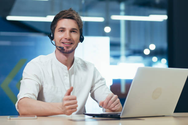 portrait of a young call center operator, a man smiling and looking at the camera, working with a laptop using a headset for a video call - hotel reception customer service representative headset receptionist imagens e fotografias de stock