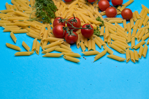 Close up shot of raw pasta tubes, cherry tomatoes and parsley on a bright blue background.