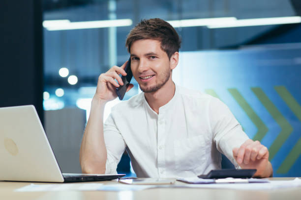 jovem bonitão, especialista trabalhando no escritório na mesa. falar em um celular com clientes, com banco, com parceiros de negócios - mobile phone business technology elegance - fotografias e filmes do acervo