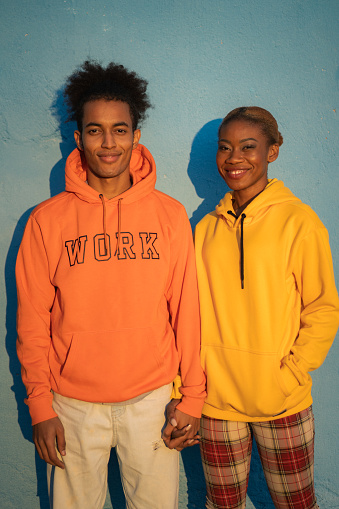 A smiling man and woman holding hands and looking at the camera wearing orange and yellow sweatshirts against a blue background