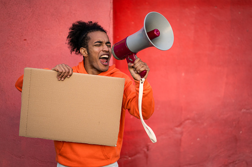 A multiethnic male activist holding a blank cardboard sign and shouting into a megaphone, wearing an orange sweatshirt against an orange wall