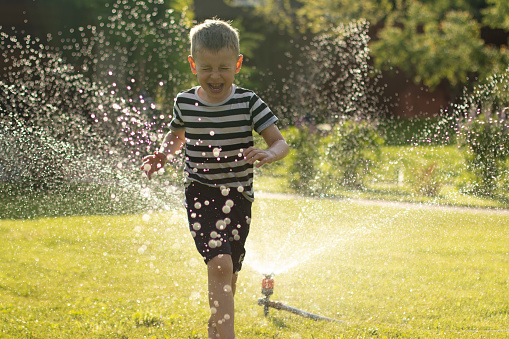 A little wet boy runs barefoot on the lawn next to the sprinkler. Happy carefree childhood and holidays concept