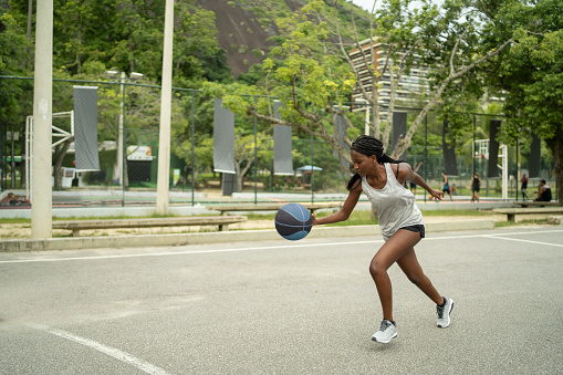 A Brazian teenage female is running and dribbling a basketball on an outside basketball court, behind her another game is in progress on a different court