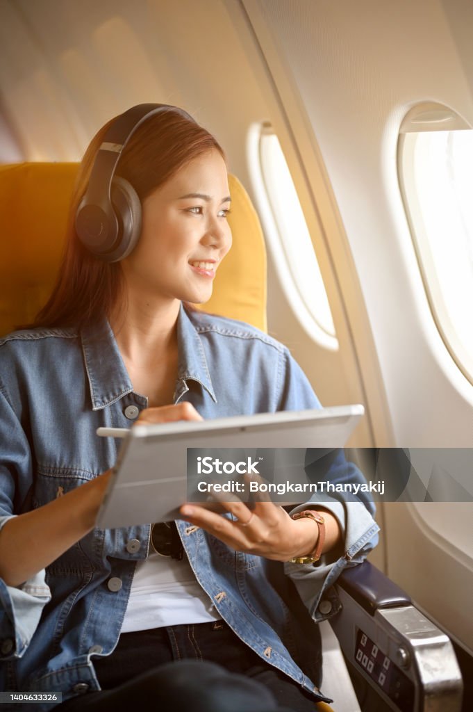 Charming asian woman passenger at window seat with headphones using portable tablet Charming millennial asian woman passenger at her window seat with her wireless headphones using portable tablet and looking out the plane's window. Airplane Stock Photo